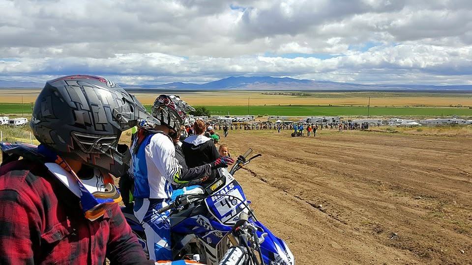 View from the ridge at Honey Lake Motocross Park: riders sitting on their bikes in the foreground, rows of RVs and trailers parked along the valley floor below, and open high-desert rangeland running out to a mountain range on the horizon.