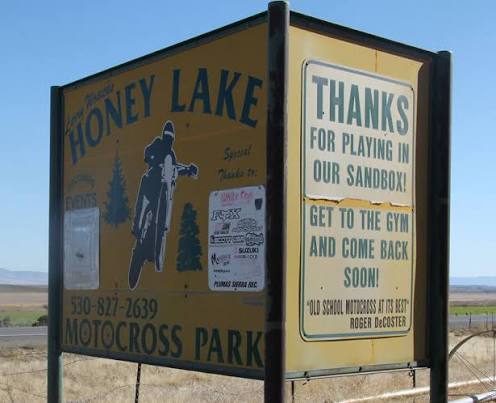 The Honey Lake Motocross Park roadside sign: a painted rider, a phone number, sponsor decals, and a panel reading "Thanks for playing in our sandbox — get to the gym and come back soon," above a Roger DeCoster quote.
